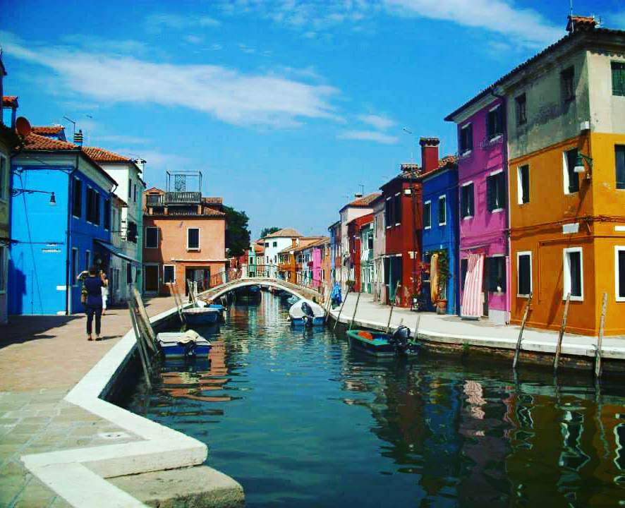 A canal with a bridge in the distance on the Venetian Island of Murano. Either side of the canal are multi-coloured brightly painted fisherman's houses