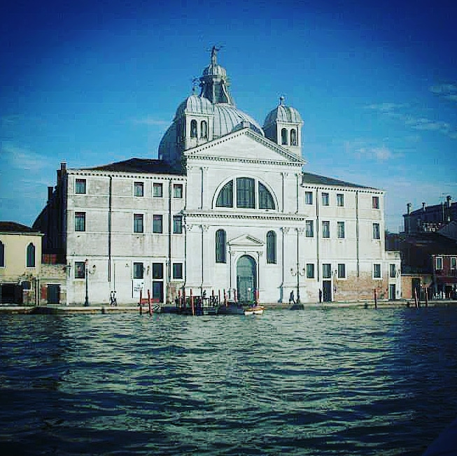 Le Zitelle Church in Venice. A large white building with large glass windows, blue door and grey domed roof. The church is situated on the edge of water