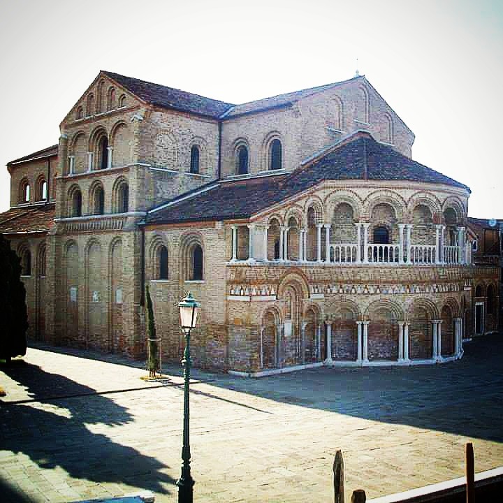 A large brown brick building with grey tiled roof. The bottom floor has pillars and doorways all along the edge and the upper floor has a balcony.