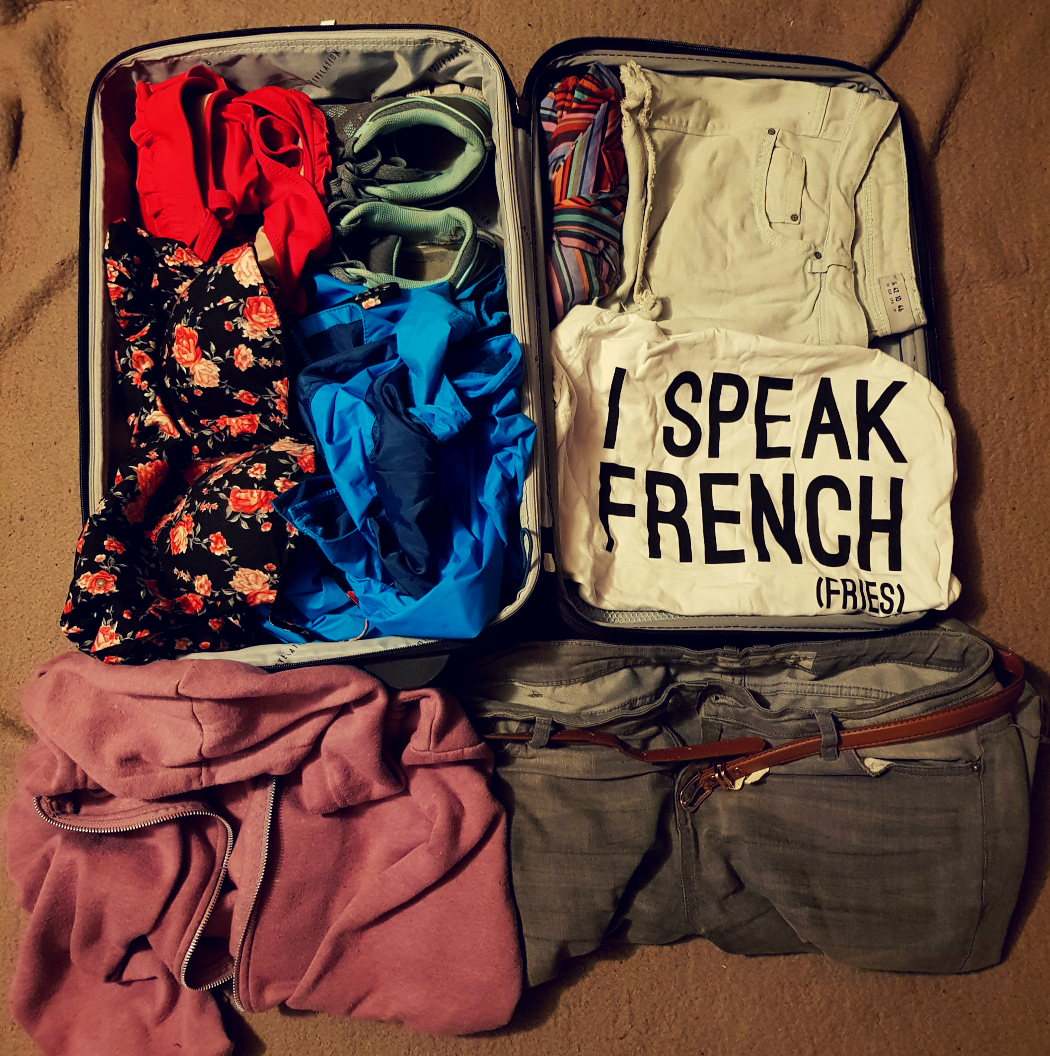 a purple suitcase filled with everything you need for your carry on bag packing in Paris France. a red bikini, blue and grey trainer, red and black flowery dress, blue raincoat, purple hoodie, grey jeans with a brown belt, blue shorts, a white top with black writing saying I SPEAK FRENCH (FRIES) and a multicoloured umbrella