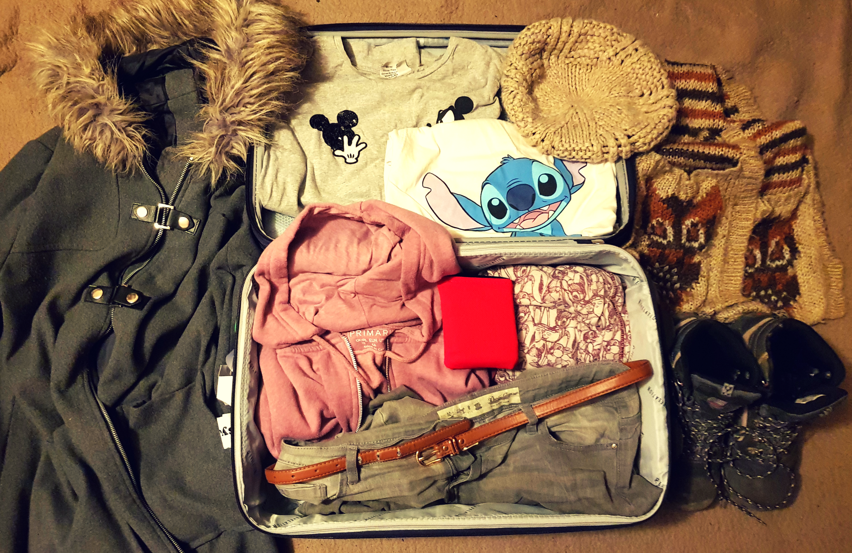 a purple suitcase with the carry on packing essentials for 3 days in France in winter. Grey winter coat with brown fur, purple hoodie, grey jeans, brown wool hat and scarf, grey and brown hiking boots, grey t-shirt with black mickey mouse heads and a blue t-shirt with Stitch from Lilo and Stitch.