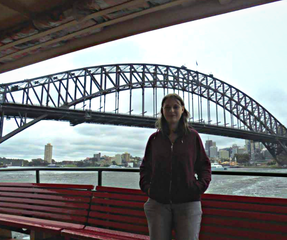A woman (me) wearing red hoodie and grey jeans stood up on a ferry with red chairs and a balcony behind. Over the balcony the Sydney harbour can be seen including the harbour bridge