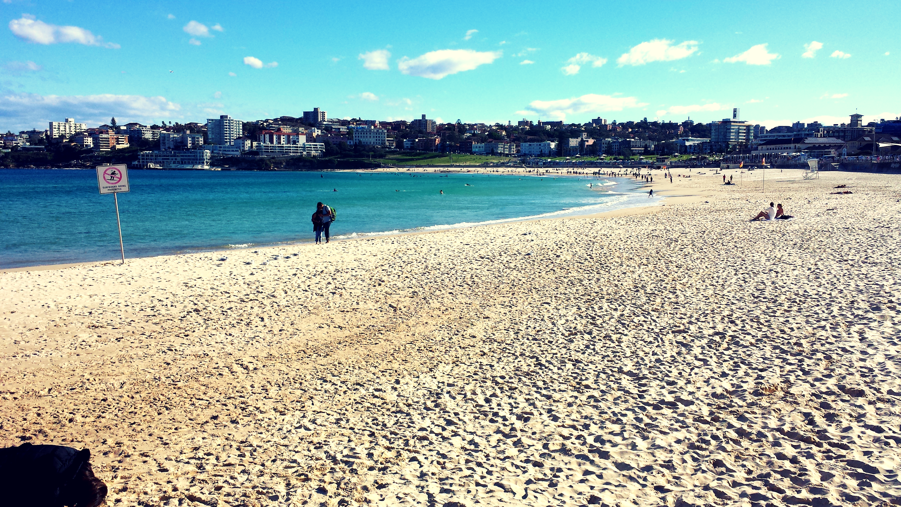 A golden sand beach stretches away from the camera in an archway with a blue sea up to the sand. In the distance is a row of buildings and houses overlooking the beach