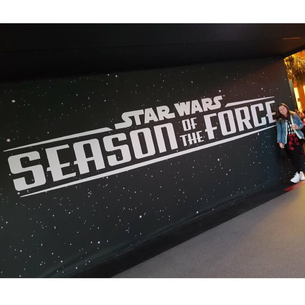 A black wall with white stars on it with Star Wars, Season of the Force written on it with a girl standing in front
