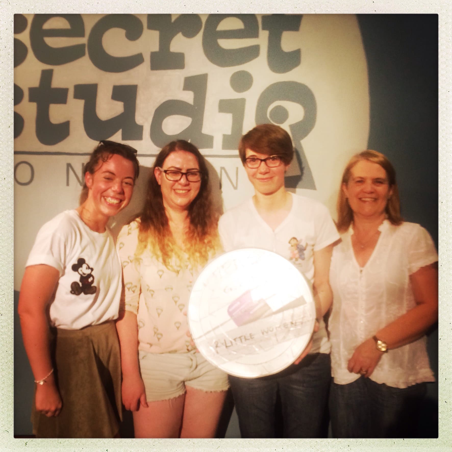 Four women stand in front of a sign saying secret studio London and holding a silver film reel with little women written on it.