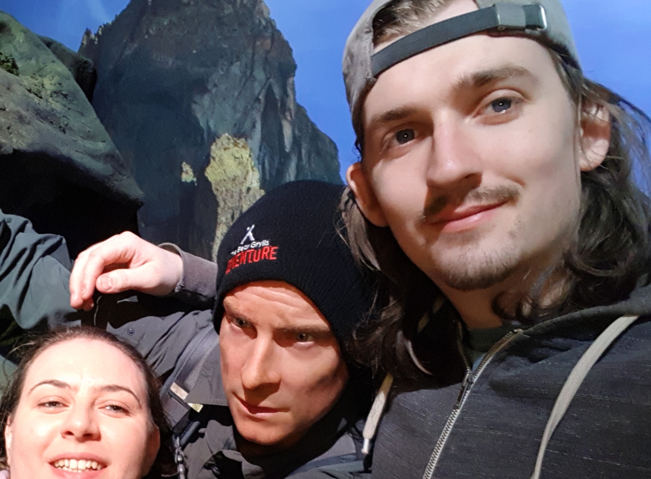 A male and female taking a selfie with a wax statue of Bear Grylls hanging off a rock