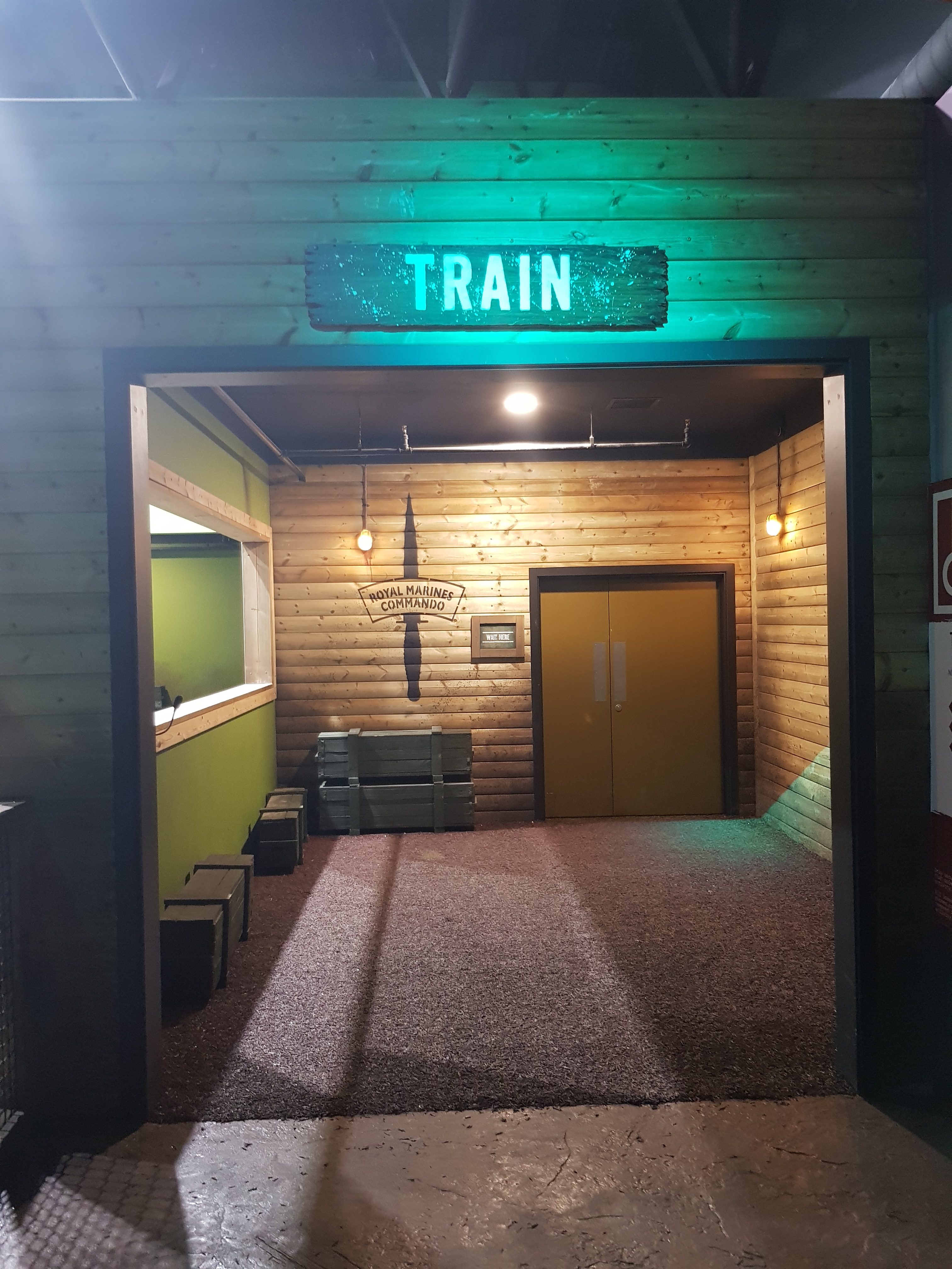 The entrance to the assault course at the Bear Grylls Adventure. A wooden door frame with TRAIN written above in white leading to a small room with a brown door and a Royal Marines Commando knife symbol.