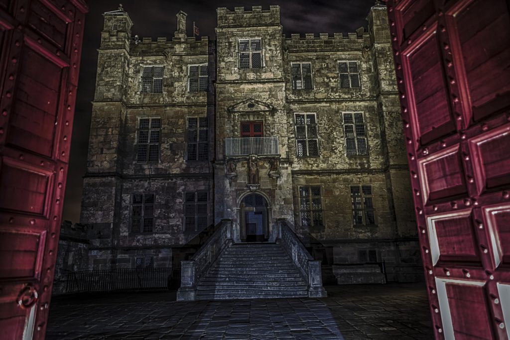 Gates opening to show Bolsover Castle. Stone steps lead up to the castle doors. A large grey stone castle with lots of windows in front of a black sky.