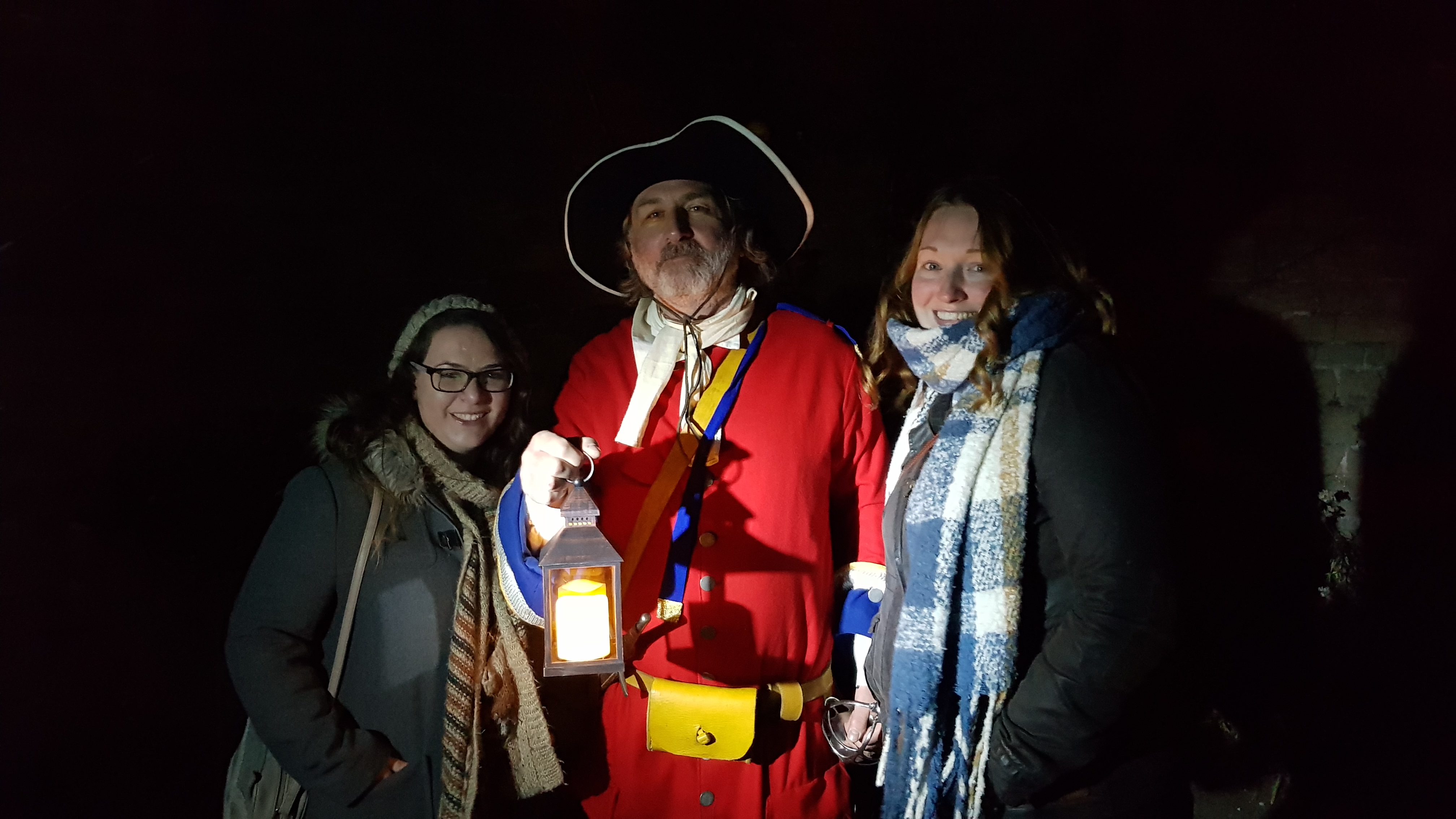 2 young girls wraped up in hats, scarf and coats smiling with an older gentlemen dressed in a red coat and black hat holding an old fashioned lantern. One of the castle guards at the Bolsover Castle Halloween fright night