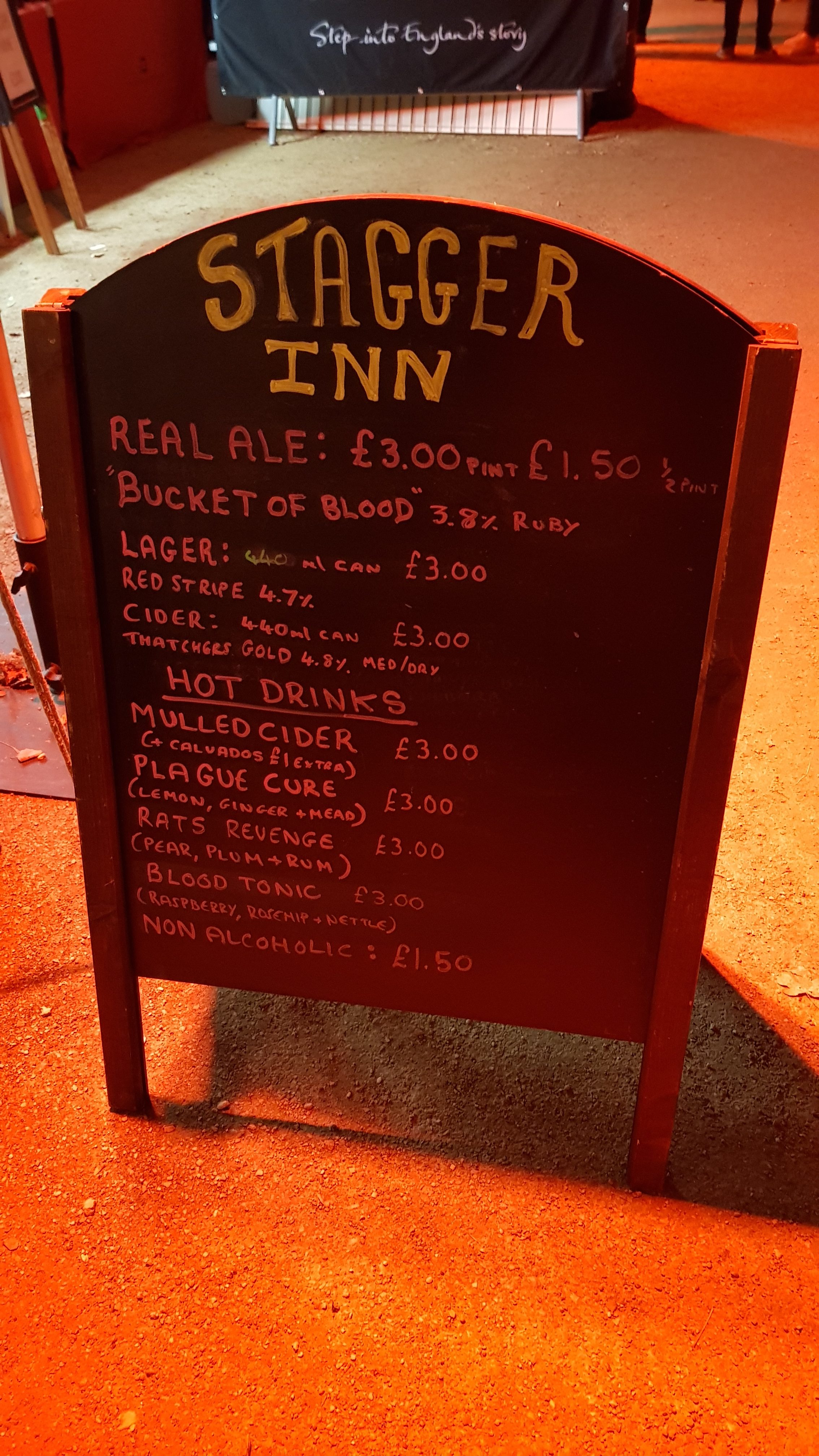 A board with the Stagger Inn menu. Real Ale, 'Bucket of Blood' £3 pint, £1.50 1/2 pint, Lager, Red Strip £3 440ml can, Cider, Thatchers Gold £3 440ml can, Hot Drinks. Mulled Cider £3, Plague Cure (lemon, ginger and mead) £3, Rats Revenge (Pear, plum and Rum) £3 Blood Tonic (Raspberry, Rosehip and Nettle) £3, Non alcoholic £1.50