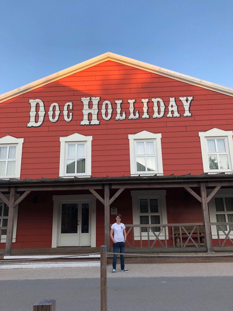 A girl standing in front of a building in the Hotel Cheyenne. A wild west style red barn type building saying Doc Holiday in white writing