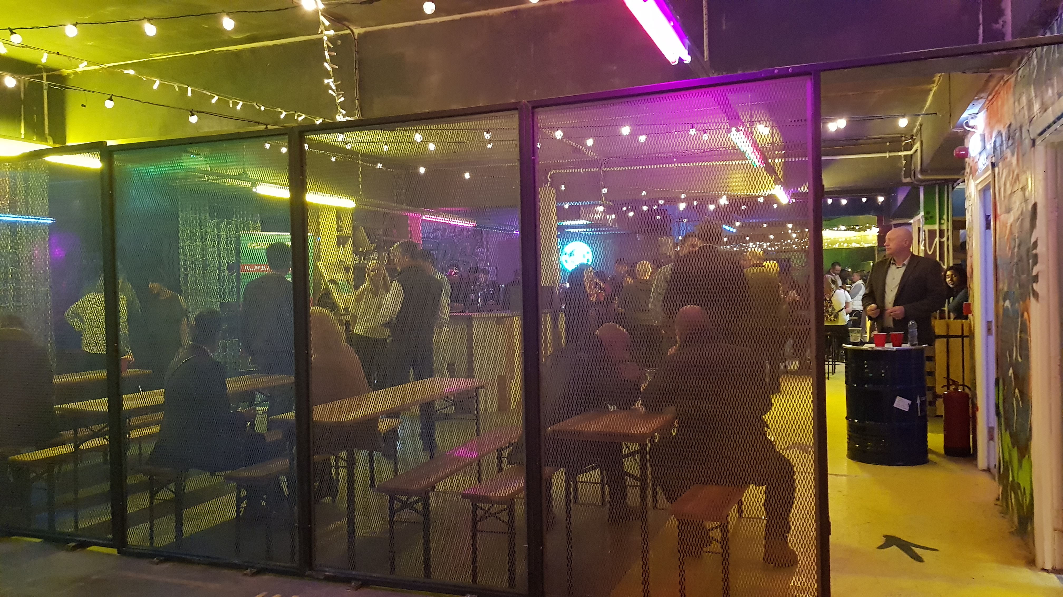 The busy bar area in Caddshackers crazy golf from behind a metal fence. A bar is in the middle of the room with wooden tables surrounding it and lots of people milling around.