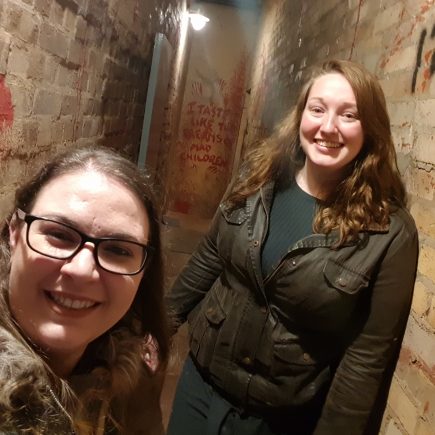 Two girls stand in a creepy looking brick corridor, with red stains and graffiti on the walls