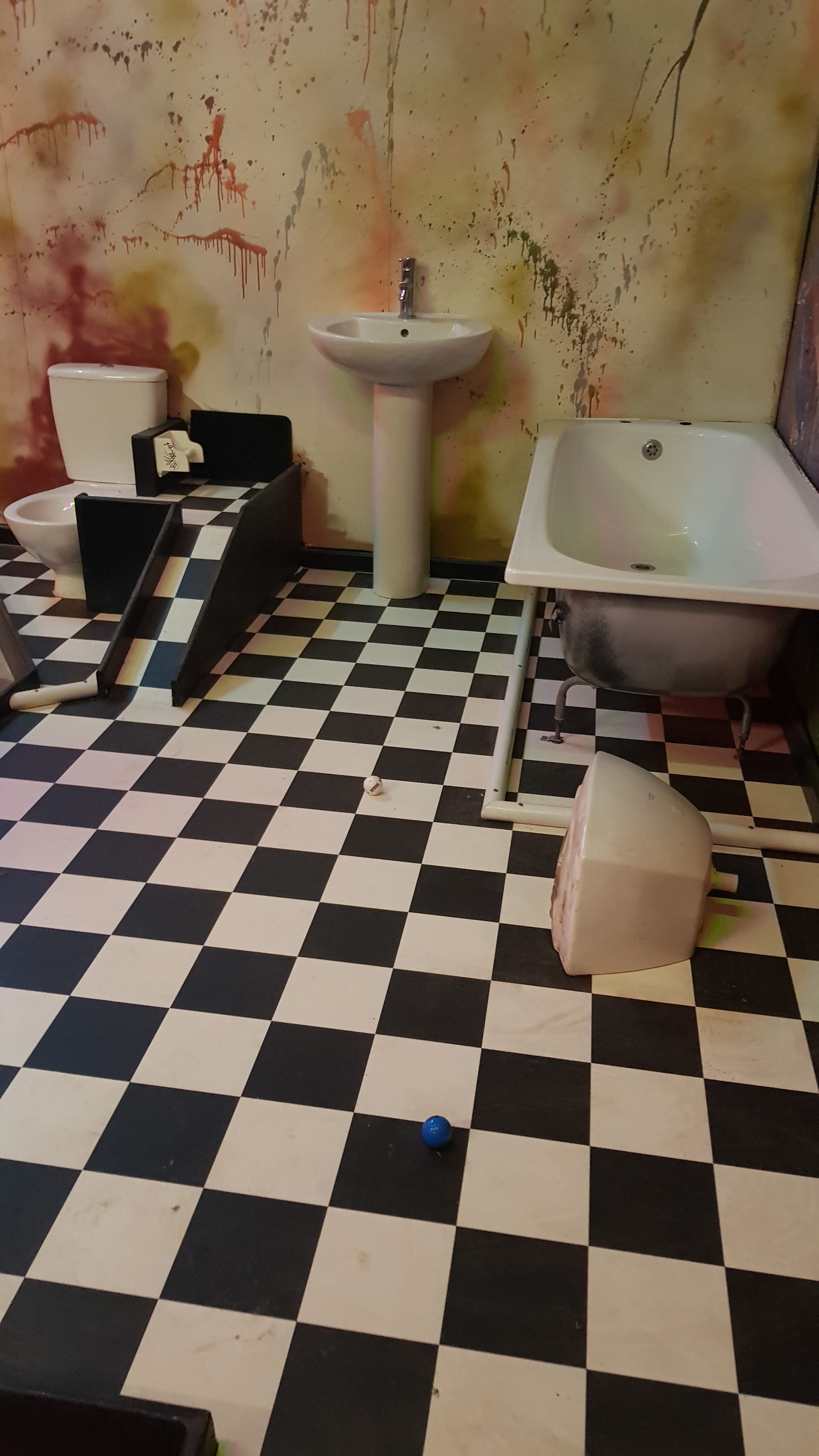 A black and white checkered floor and cream walls covered in stains. A bath , sink and toilet are in the room with a ramp going up next to the toilet to get the ball into the hole which is built into the toilet.