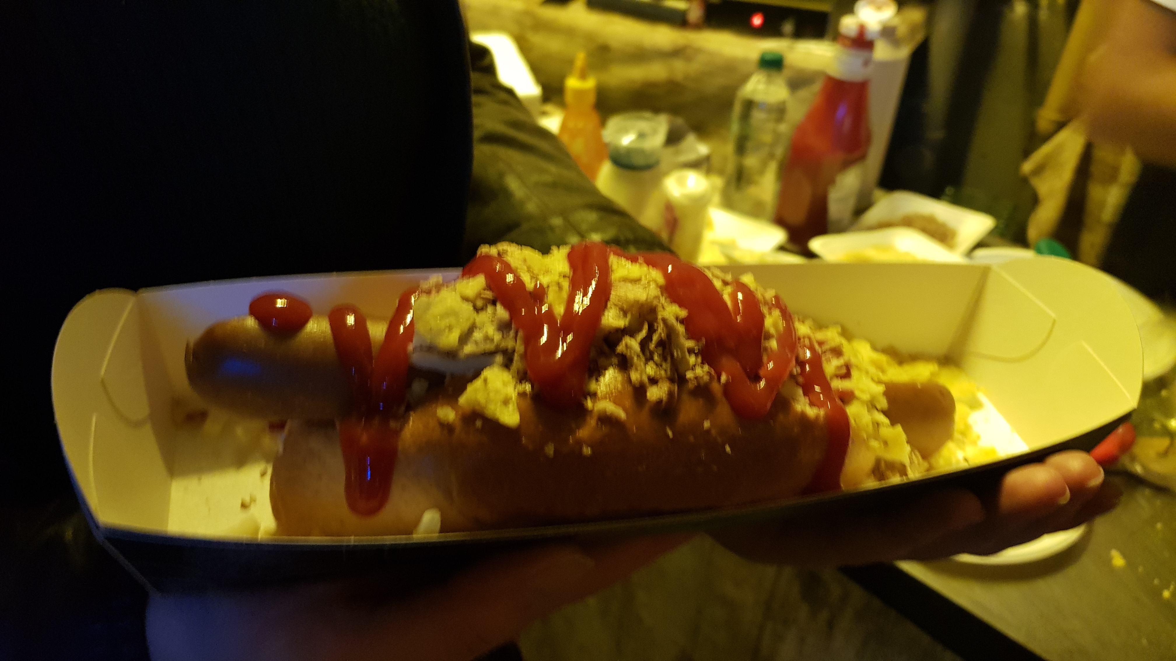 A vegan sausage in a hotdog bun, covered in onions, nachos, vegan cheese and tomato ketchup