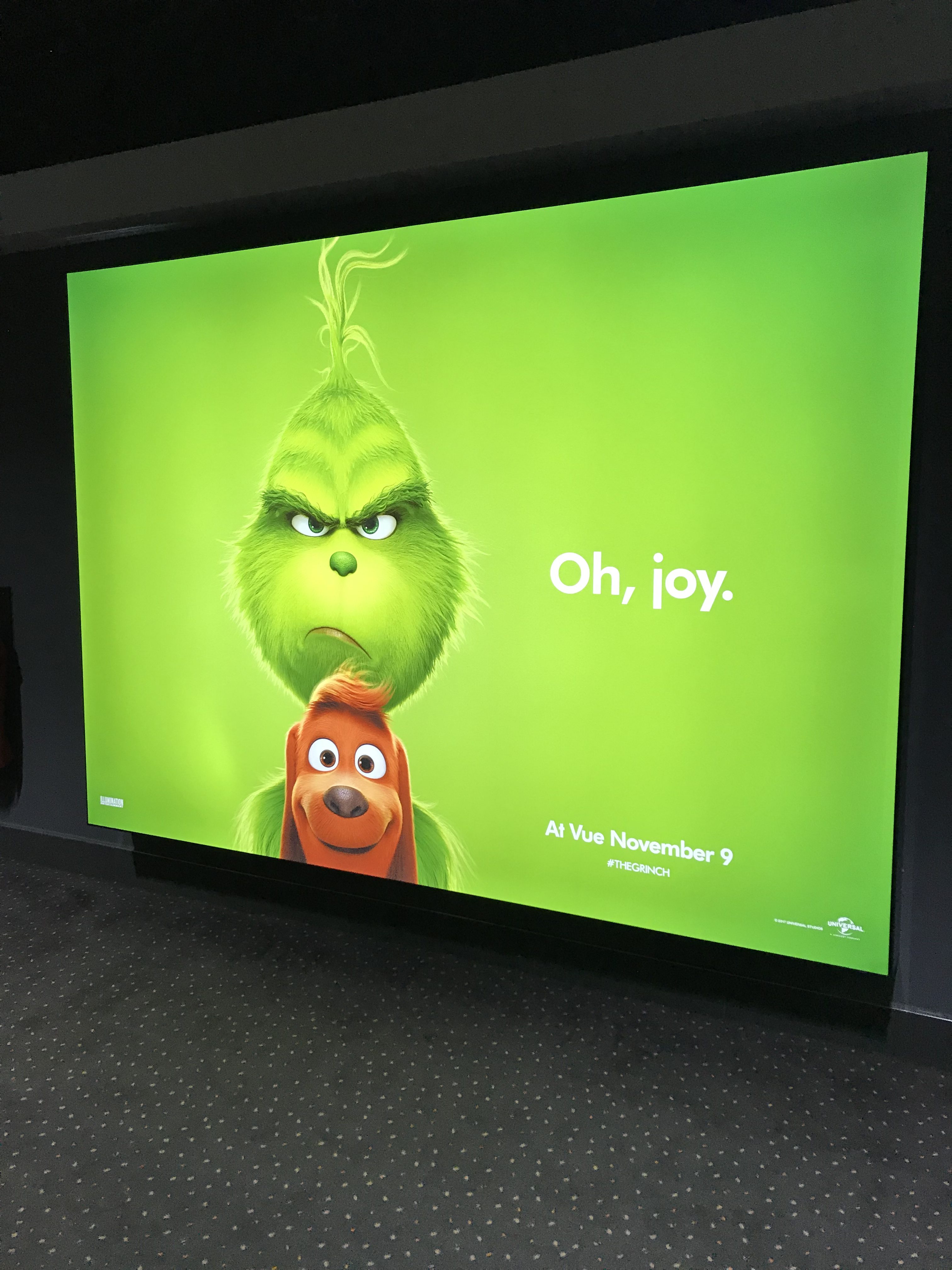 A poster for the Grinch Christmas film. The background is green with a cartoon version of the Grinch, (a green furry creature with an angry expression) holding a brown dog. Next to the Grinch it says 'Oh Joy.'