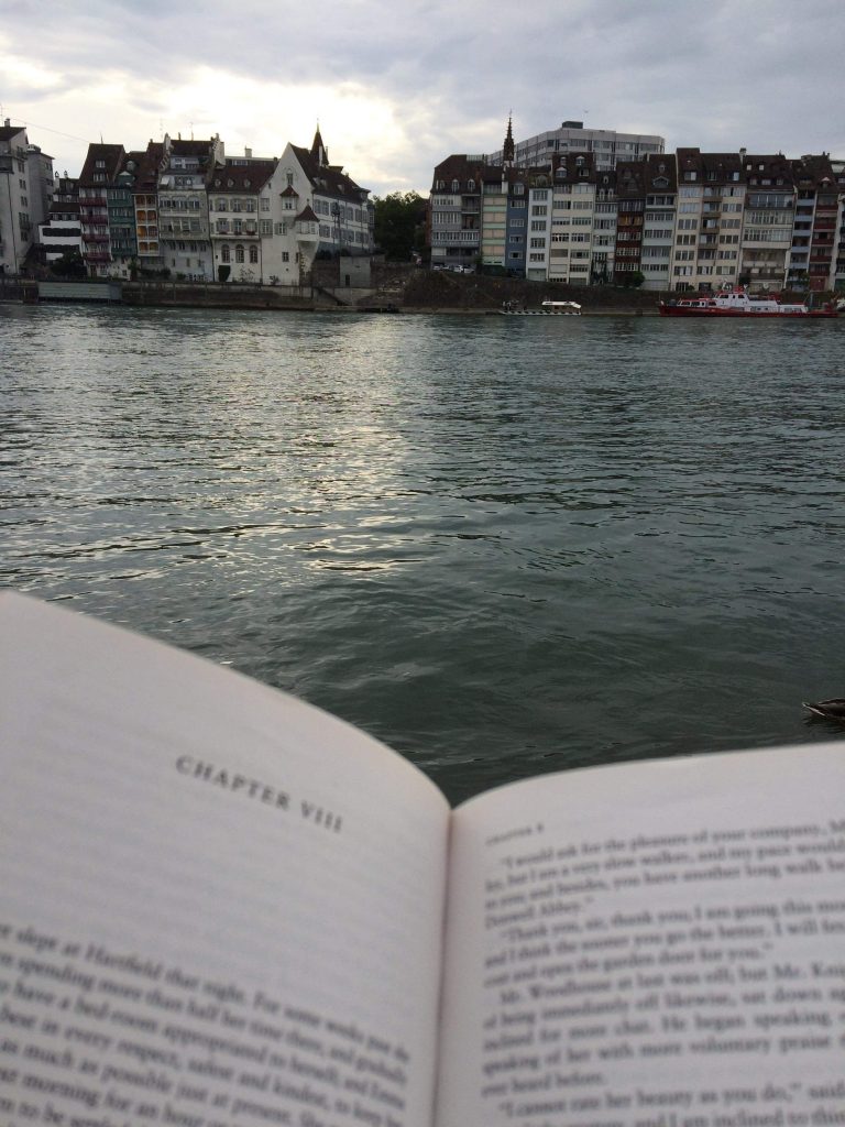 Reading a book while sat on the edge of the river Rhine with buildings on the other bank