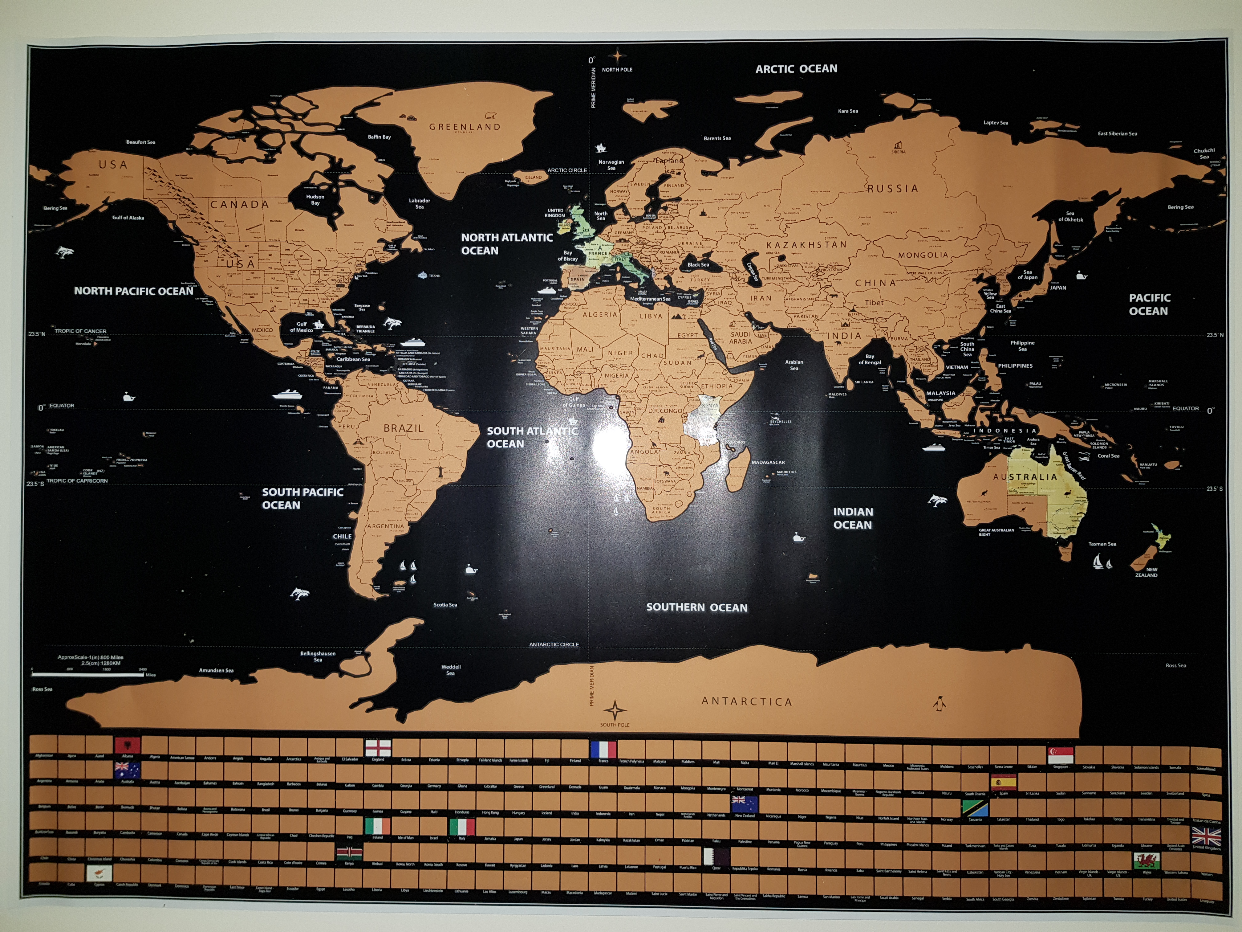 A map of the world where the water is black and the land is gold foil which can be scratched off. At the bottom of the map is scratch off boxes covering the flags of every country