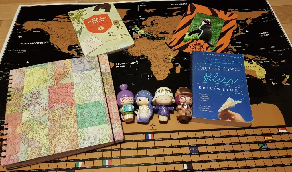 A few travel items that make great gifts for travelers. A black and gold scratch off world map, a scrapbook with a map of the world, a book called the Small Adventure Journal, a book called The Geography of Bliss, a picture of a penguin in a Dudley Zoo tiger striped frame and four momiji dolls, a traveler holding a map, a mermaid, a girl wearing a star dress and headband and a detective doll.