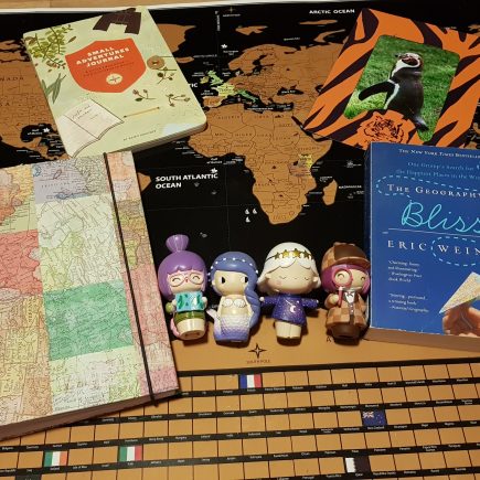 A few travel items that make great gifts for travelers. A black and gold scratch off world map, a scrapbook with a map of the world, a book called the Small Adventure Journal, a book called The Geography of Bliss, a picture of a penguin in a Dudley Zoo tiger striped frame and four momiji dolls, a traveler holding a map, a mermaid, a girl wearing a star dress and headband and a detective doll.