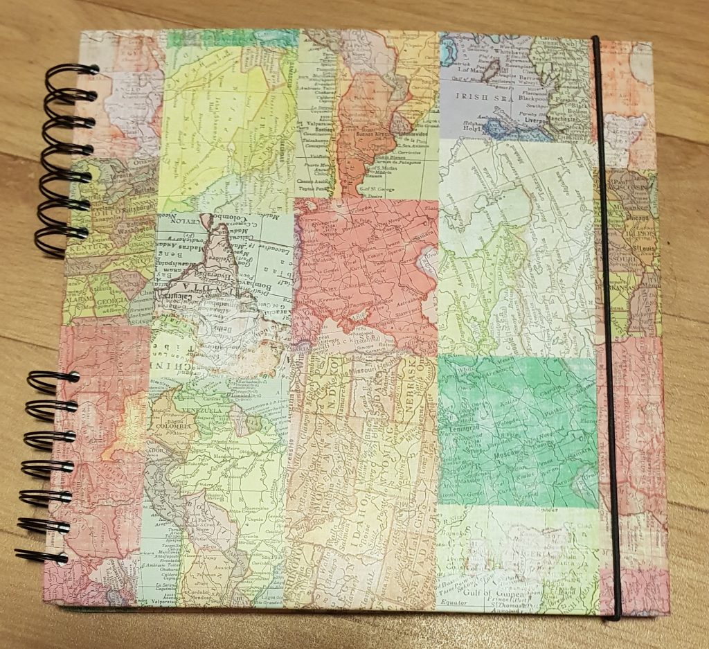 A scrapbook with multicoloured sections of different maps in a patchwork pattern on the cover and a black elastic string holding it shut.