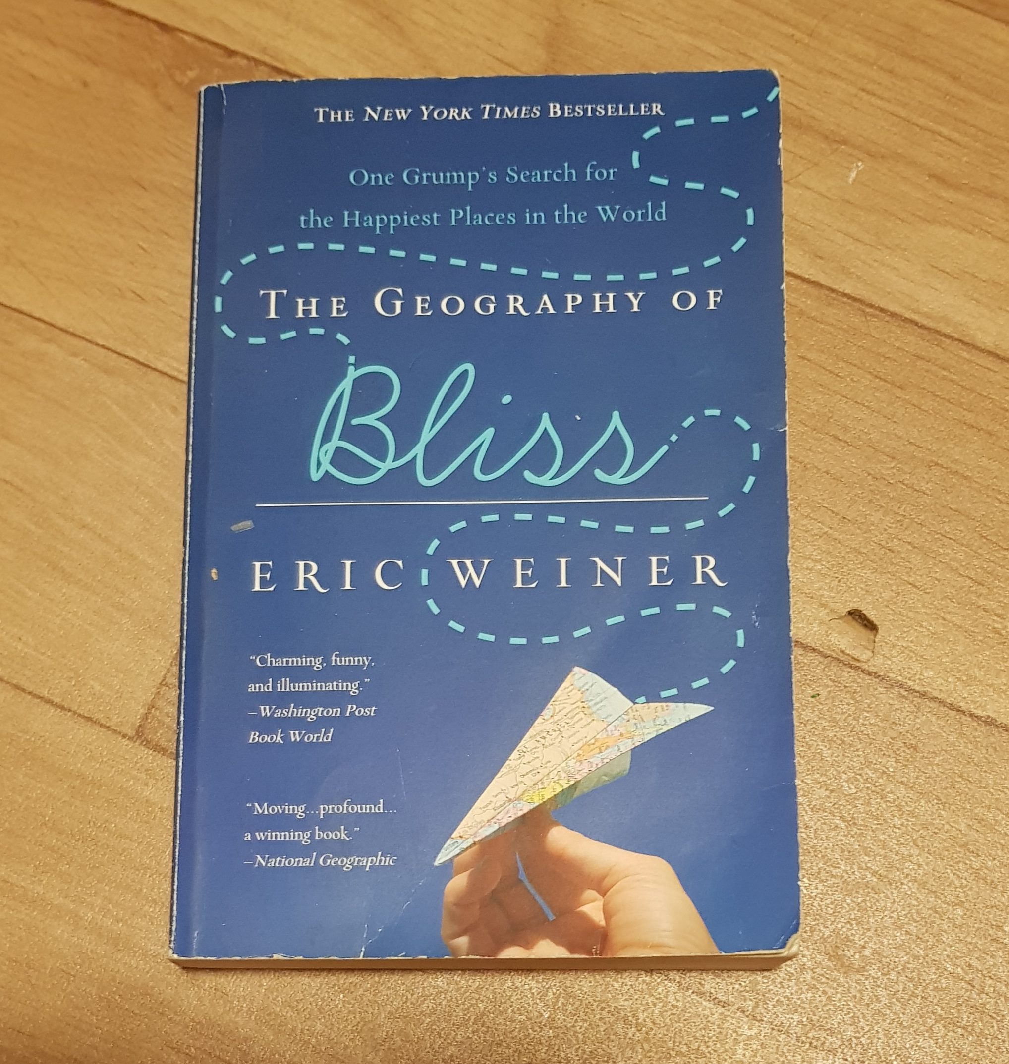 A blue book with a hand holding a paper aeroplane. The writing says The Geography of Bliss, one Grump's search for the happiest places in the World