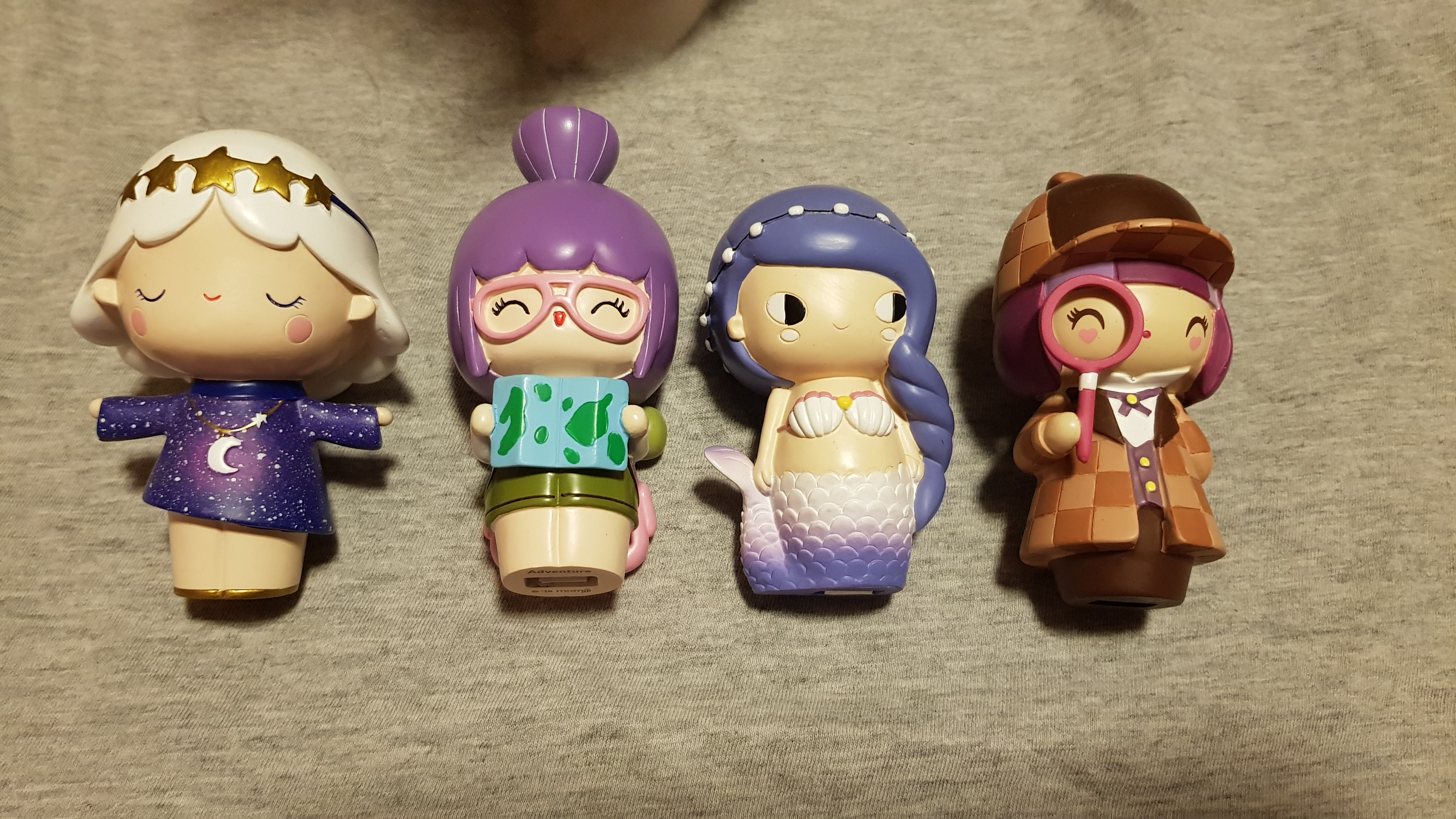 Four small dolls made of tree resin. The first has white hair with a gold star headband and blue dress with white stars on it and a moon necklace. The second has purple hair and glasses and is holding a map of the world. The third has a purple mermaid tail, shell bra, purple hair and a headband. The fourth is wearing a brown checkered coat and detective cap and is holding a pink magnifying glass.