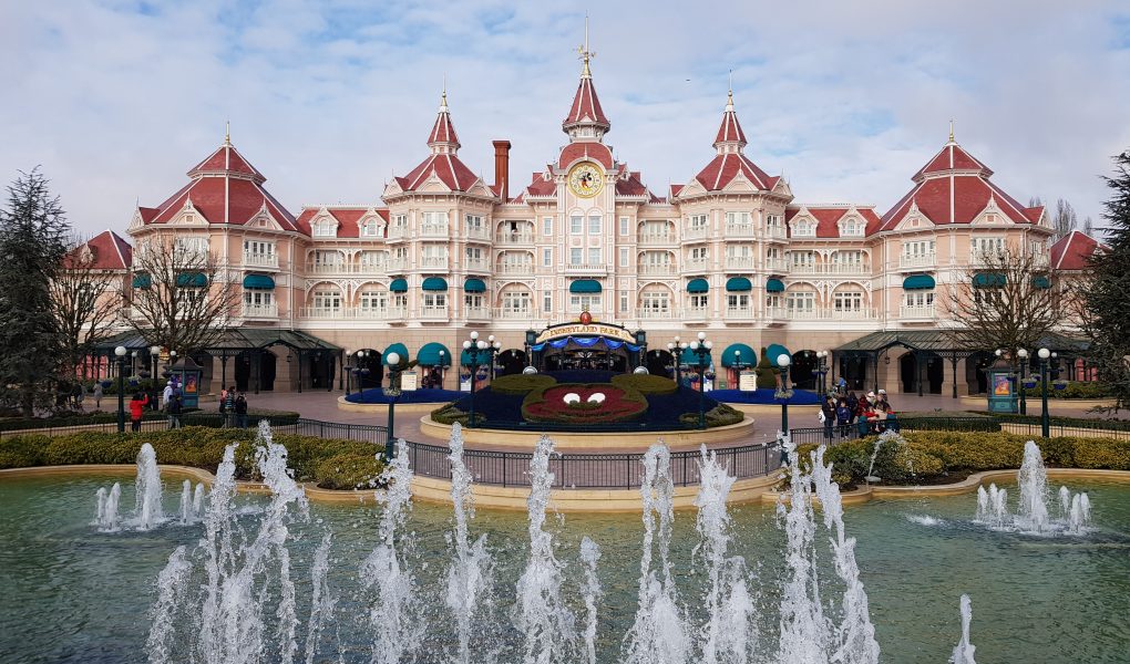 The Disneyland Hotel at Disneyland Paris. A pale pink castle shaped building with dark pink roof. A red mickey mouse head is made out of flowers in a circular flower bed and a small lake with fountains shooting water upwards was in front of the building