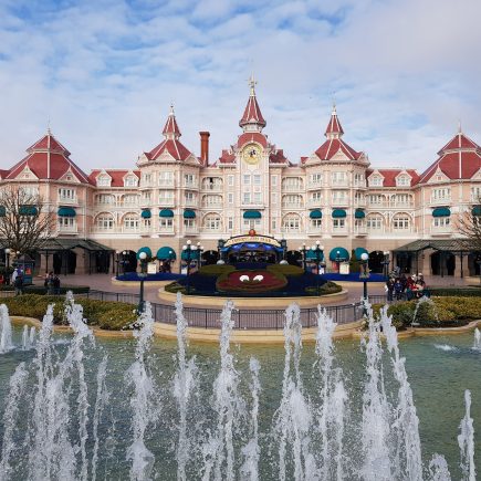 The Disneyland Hotel at Disneyland Paris. A pale pink castle shaped building with dark pink roof. A red mickey mouse head is made out of flowers in a circular flower bed and a small lake with fountains shooting water upwards was in front of the building