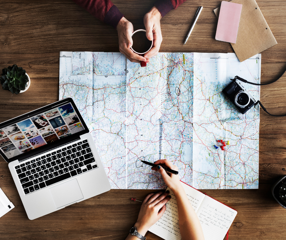 A flatlay image of two people starting their plans on their travel goals. A map is laid out over the table with an open laptop, camera and two sets of arms pointing out areas on the map.