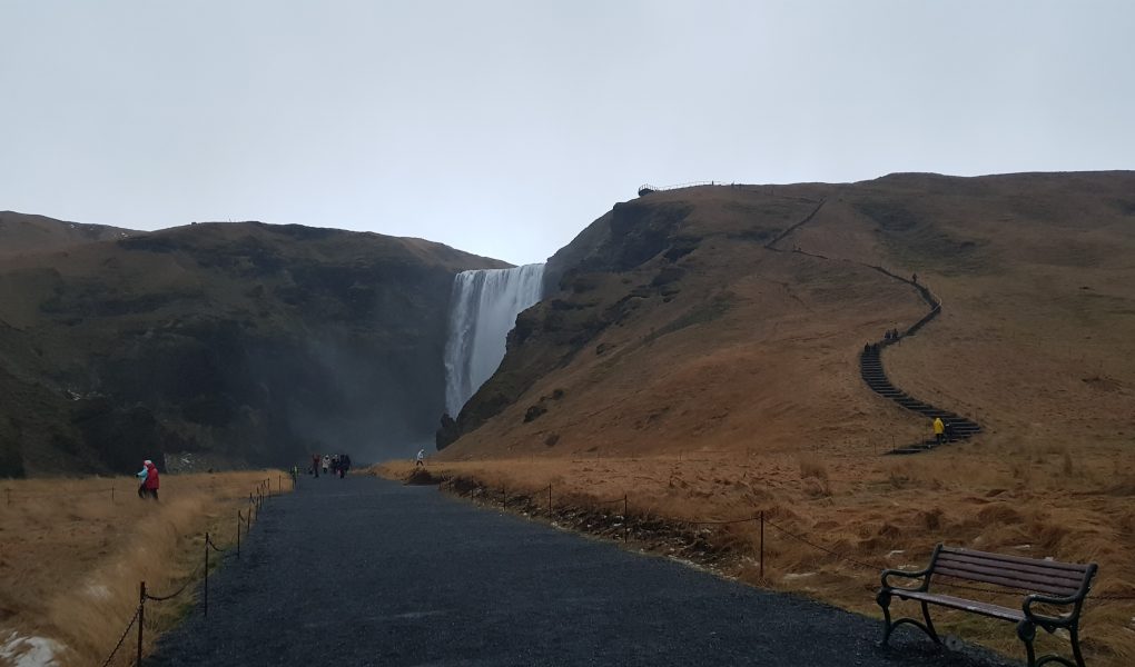 A wide dark path leads away from the camera and up to a waterfall in the distance. Either side of the waterfall and large hills and one side has a very steep pathway leading up the hill