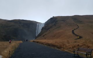 A wide dark path leads away from the camera and up to a waterfall in the distance. Either side of the waterfall and large hills and one side has a very steep pathway leading up the hill