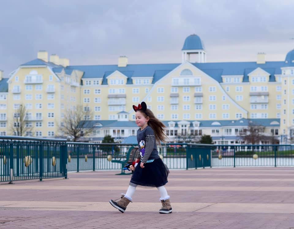 A little girl wearing Minnie Mouse ears outside the Newport Bay Club hotel. A pale yellow building with a blue roof