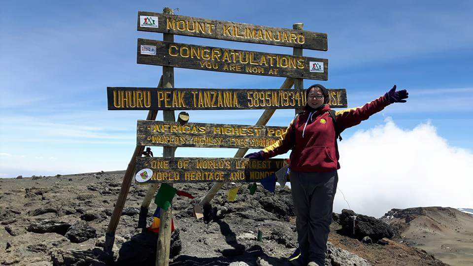 A girl stood at the summit of Kilimanjaro. A sign reads 'Mount Kilimanjaro. Congratulations you are now at Uhuru Peak 5895m'