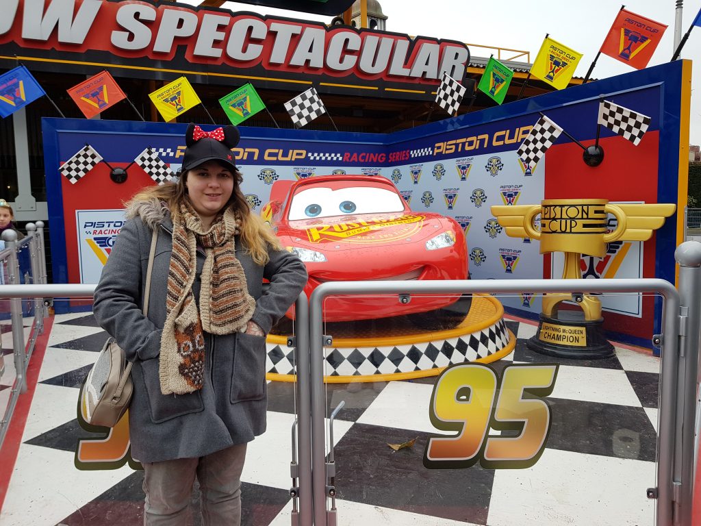 A girl stood in front of a car designed to look like lightning Mcqueen with red paint and eyes instead of a window. Next to him stands a large trophy with Piston Cup written on it