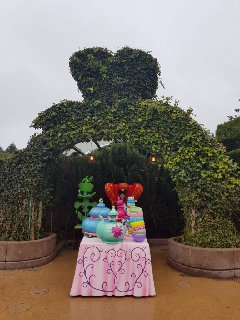 Arch way made out of shrubbery with a heart on the top. Inside the archway is a table laid out with tea pots and cups with the Dormouse peeping out of a teapot