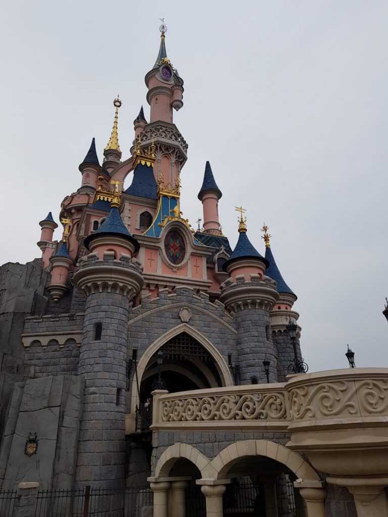 A shot of Sleeping Beauty's Castle at Disneyland Paris. A pink castle with blue roofs with gold details and a round stained glass window. A bridge leads into the main doorway through a grey stone section