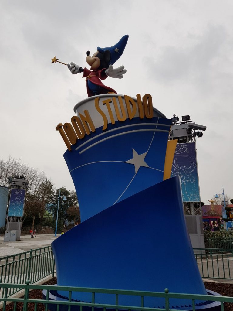 Statue of Mickey Mouse in wizards outfit from Fantasia. He reaches into the air with a yellow star wand, has a red gown on and blue starry wizards hat. He stands on a blue platform with Toon Studio written in gold.