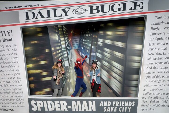 two girls posing like they are firing webs out their hands with spiderman dressed in red and black full body suit and a background shows a newspaper page of the Daily Bugle