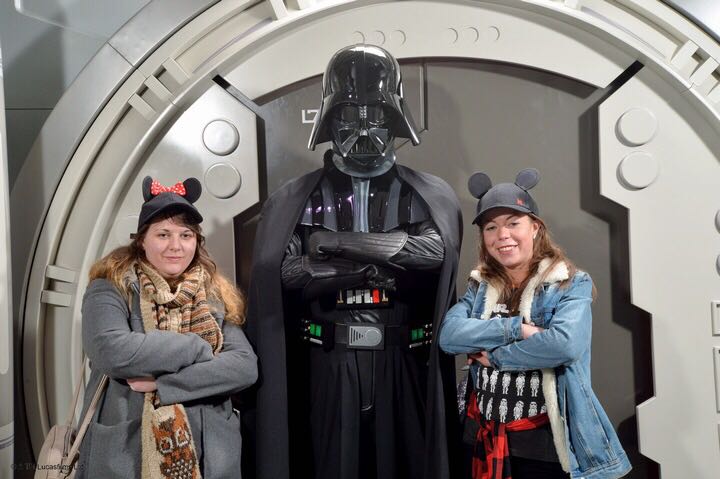 2 girls wearing Mickey ear hats stood next to darth vader in black outfit and helmet. All three are crossing their arms and looking stern