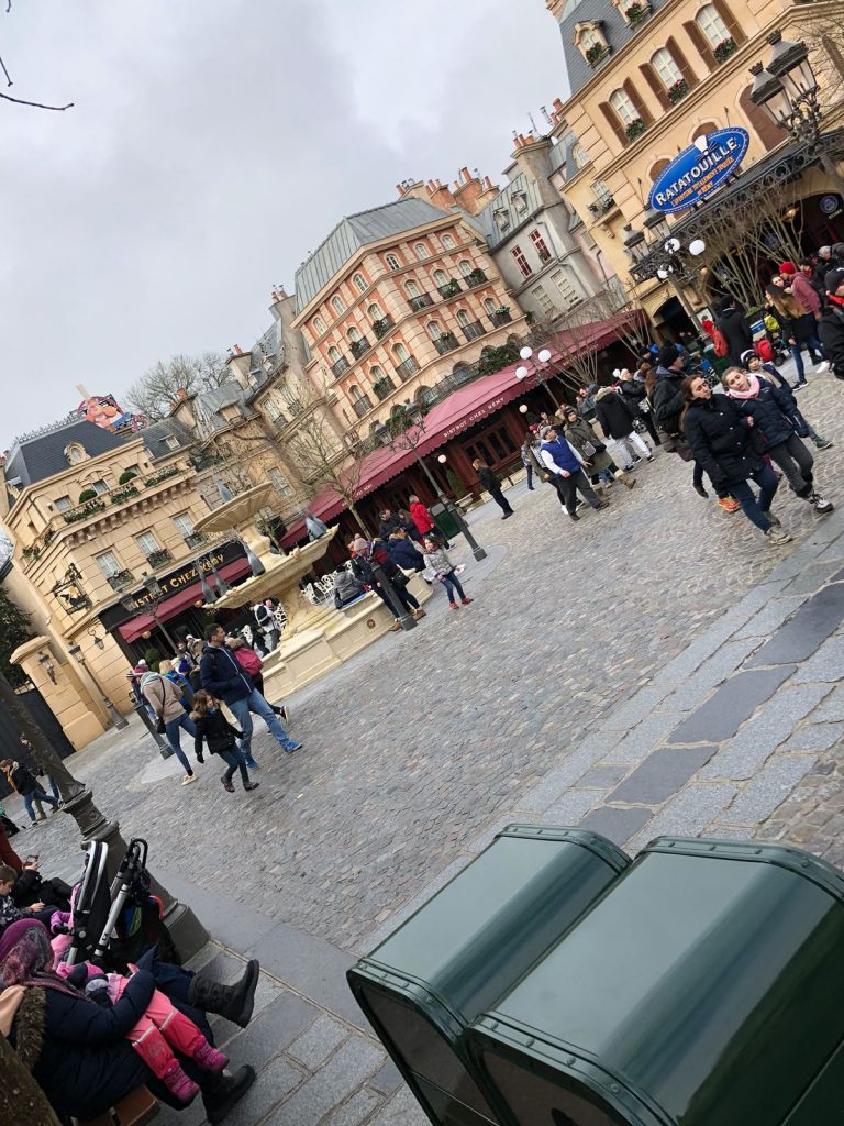 Parisian street scene in front of the Ratatouille ride. A fountain is in the middle of the square with tall buildings around the edge. A restaurant has red awning over the ground floor of the building
