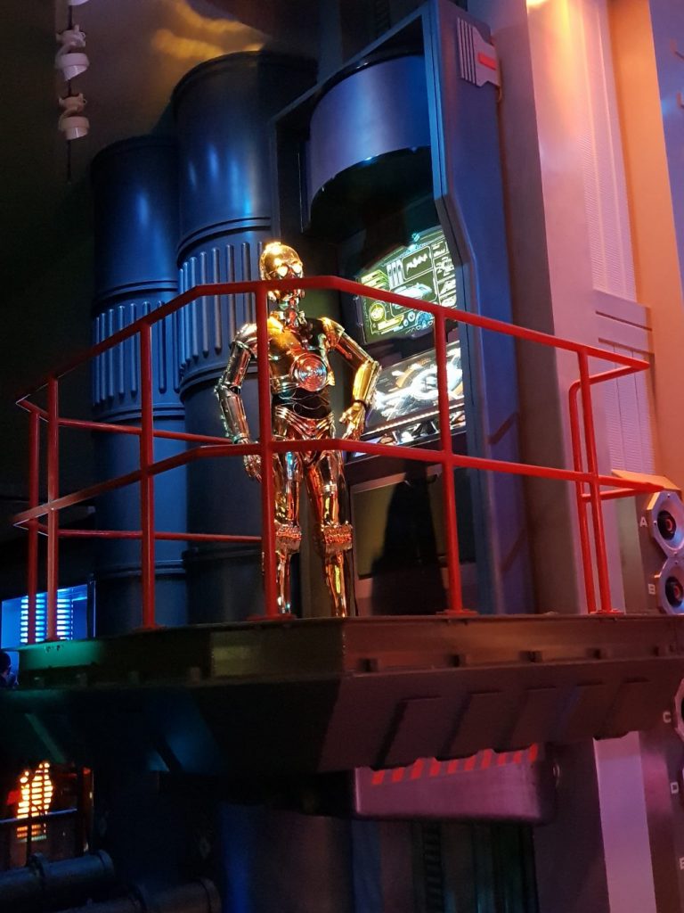C-3PO a gold metal robot found inside th Star Wars Hyperspace Mountain. He is standing on a platform in front of a computer