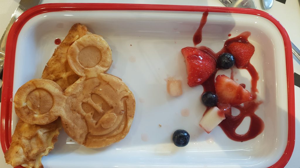 A waffle shaped like a Mickey Mouse head sat on a long waffle next to some fruit in raspberry compote on a white tray with red border. These are found in the Disney cafe at the world's largest Primark in Birmingham.