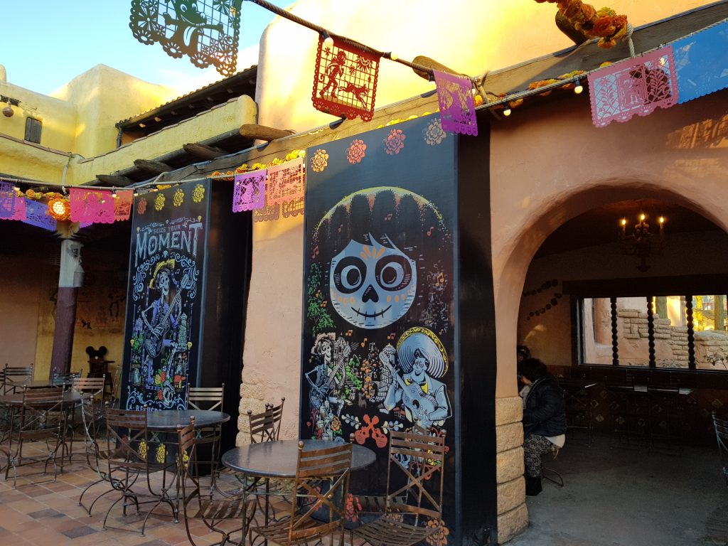 The inside of a courtyard at Fuente del Oro Restaurante. Orange archways line the walls. Colourful flags with cut out shapes are strung up along the walls and across  the middle of the courtyard. Two black panels have colourful pictures of Mexican style sugar skull skeletons.