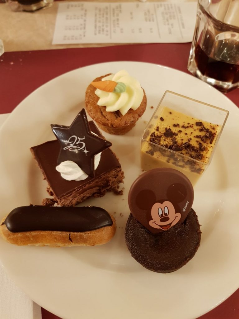 Desserts on a plate. A mini Chocolate eclair, a mini chocolate cake with a chocolate circle with Mickey mouse printed on it, a small cholate square cake with a chocolate star saying 25, a mini cheese cake with caramel top in a glass and a mini carrot cake with some butter cream and a carrot decoration.