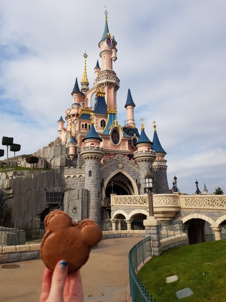 a hand holding a chocolate macaroon in the shape of Mickey's head. Behind is the Disneyland Paris Castle with grey stone walls, a cream bridge, pink turrets and blue roofs 