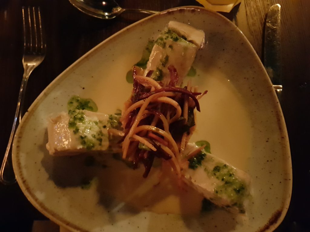 A plate with three fillets of white fish in a garlic sauce. In the middle is a pile of mash potato covered in shredded carrots that are orange and purple