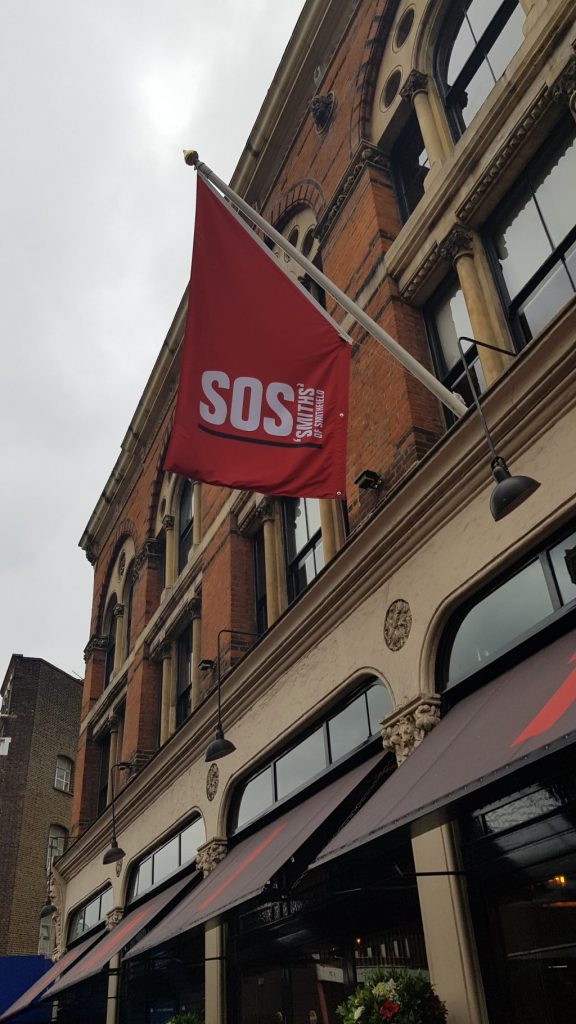A tall building with a restaurant on the ground floor and black awning. A red flag is hung with white writing saying SOS Smith's of Smithfield. This is the starting location for Lost Souls of the City