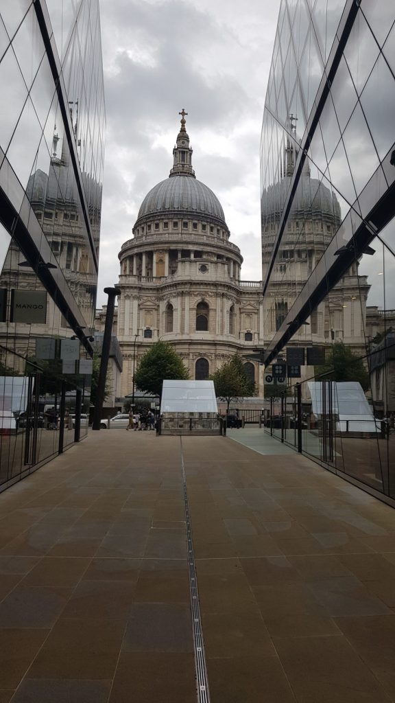 Photograph of St Paul's Cathedral taken between two large glass buildings so that the cathedral reflects against them. The Cathedral is limestone with arches, pillars and a large domed roof