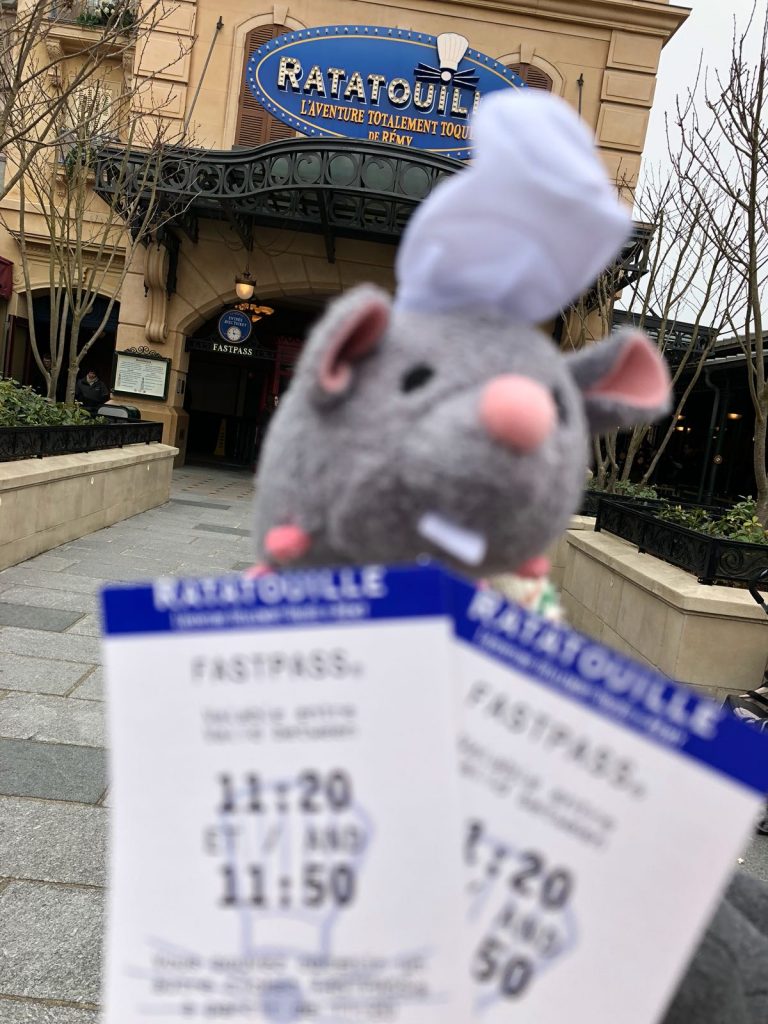 Two Disneyland Paris fastpass tickets for the Ratatouille ride in front ot the door to the ride with its title above. A stuffed mouse wearing a chef house is held above the tickets